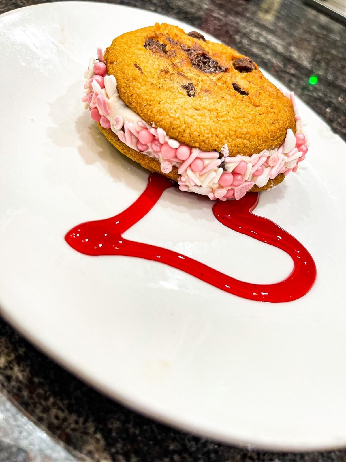 A chocolate chip cookie sandwich with pink and white sprinkles, served on a plate with a red heart-shaped sauce garnish.