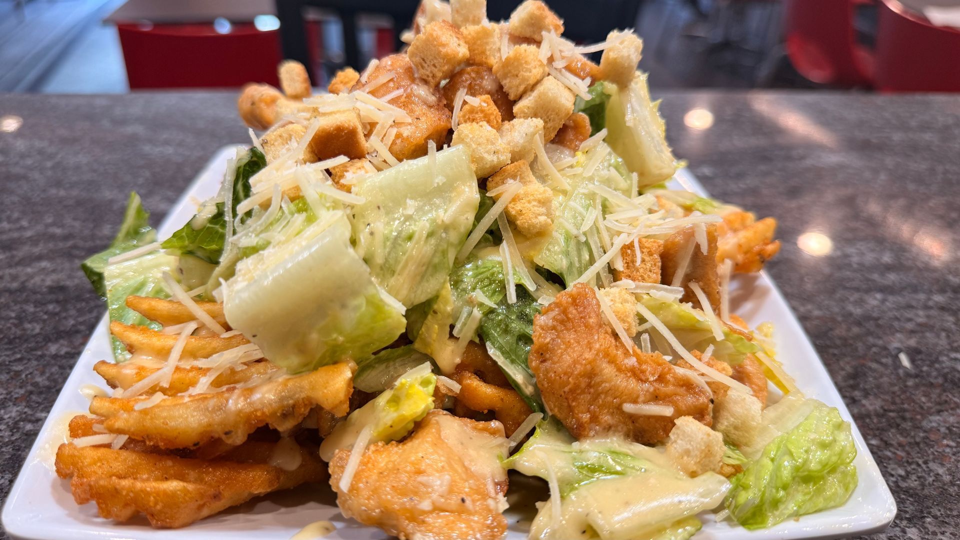 A Caesar salad with fried chicken and waffle fries on a white rectangular plate sitting on a dark stone countertop.