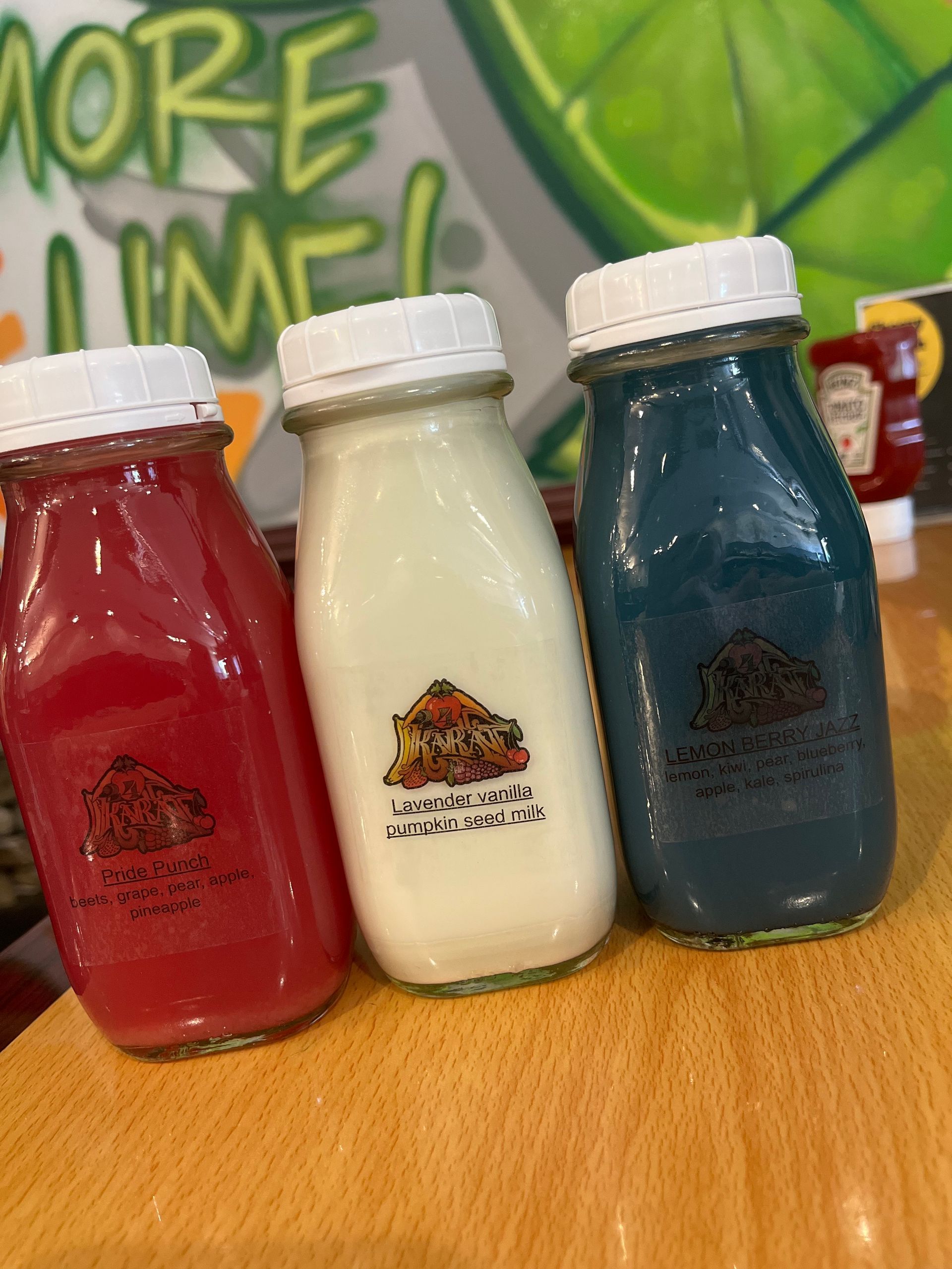 Three glass bottles filled with red, white, and blue drinks sit on a wooden table in front of a lime-themed wall graphic.