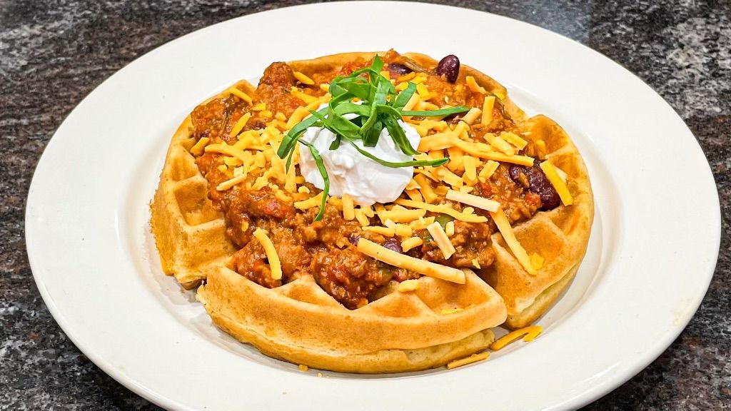 A golden-brown waffle topped with chili, shredded cheddar cheese, a dollop of sour cream, and fresh green herbs.