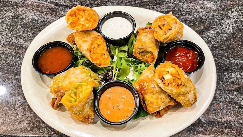 A plate of crispy, golden-brown egg rolls arranged on a bed of greens with three small cups of colorful dipping sauces.