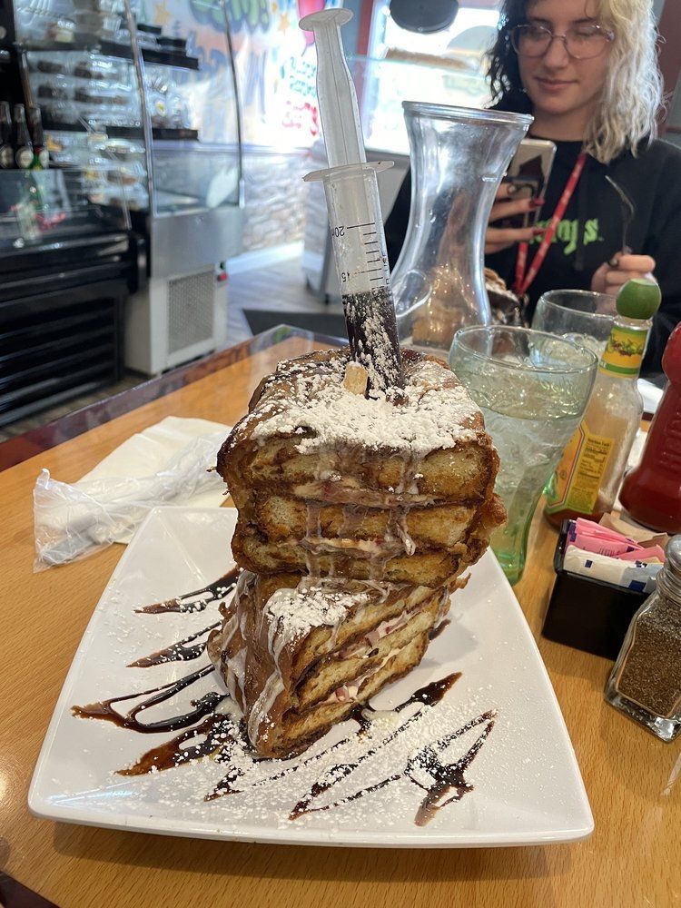 Tall stack of French toast, syrup drizzled, powdered sugar, syringe filled with cream, person in background.