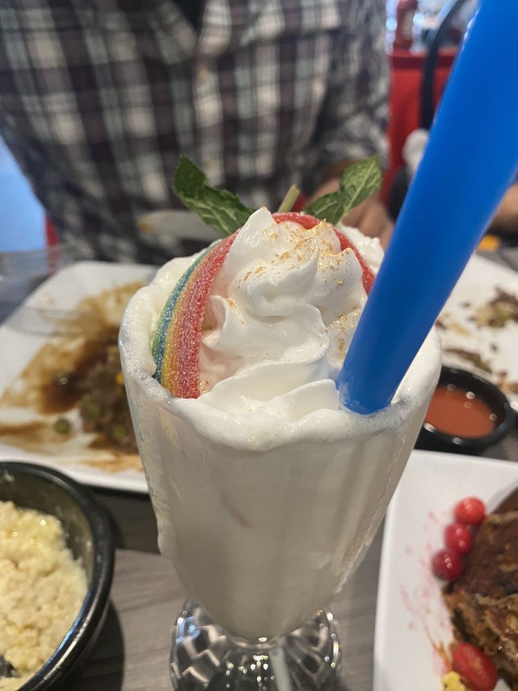 Milkshake with whipped cream, rainbow candy, mint, and blue straw, at a restaurant.