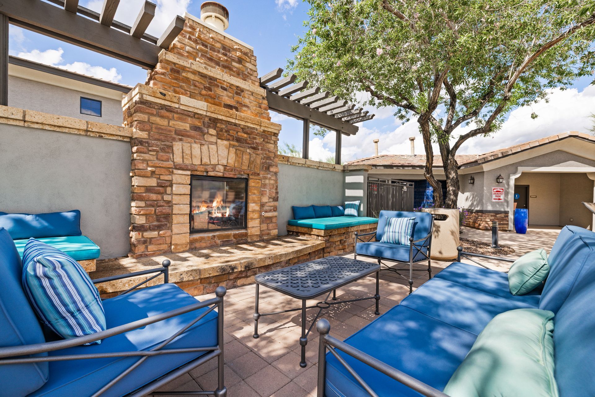 Backyard patio with blue seating around a brick fireplace under a pergola, beside a tree and house.