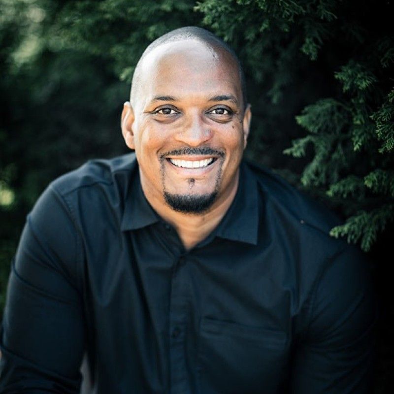 Man with shaved head and goatee smiles broadly, wearing a black shirt, set against green foliage.