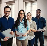 Four smiling colleagues in an office, holding papers and a laptop.