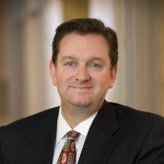 Man in suit smiles, indoors, possibly a business setting.