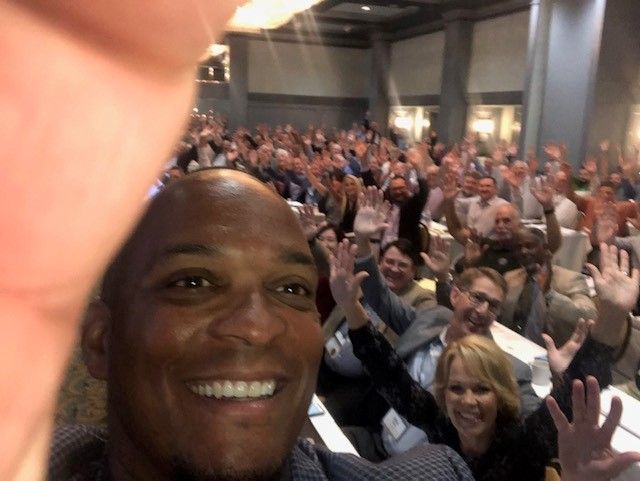Person writing in a notebook at a table with a laptop and another person using a phone.