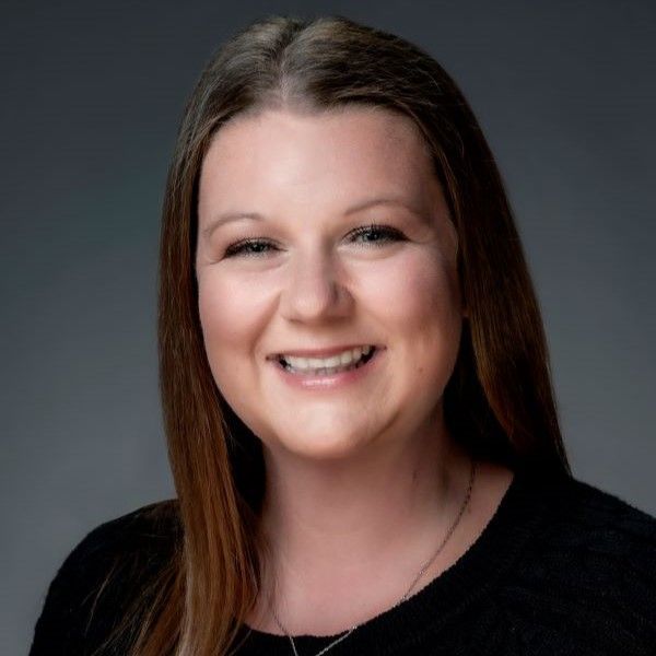 Woman with long, light-brown hair smiles at the camera, wearing a black top against a grey background.