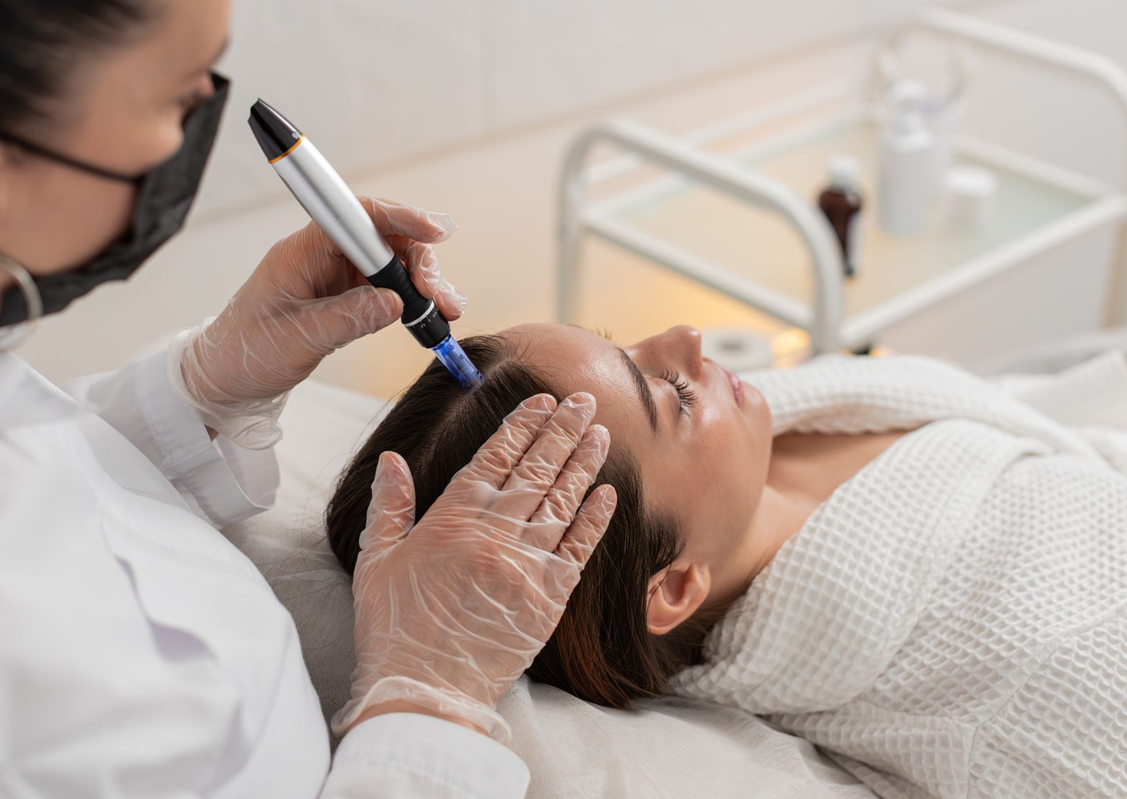 A practitioner in a white coat and gloves uses a microneedling device on a person’s scalp in a clinical setting.