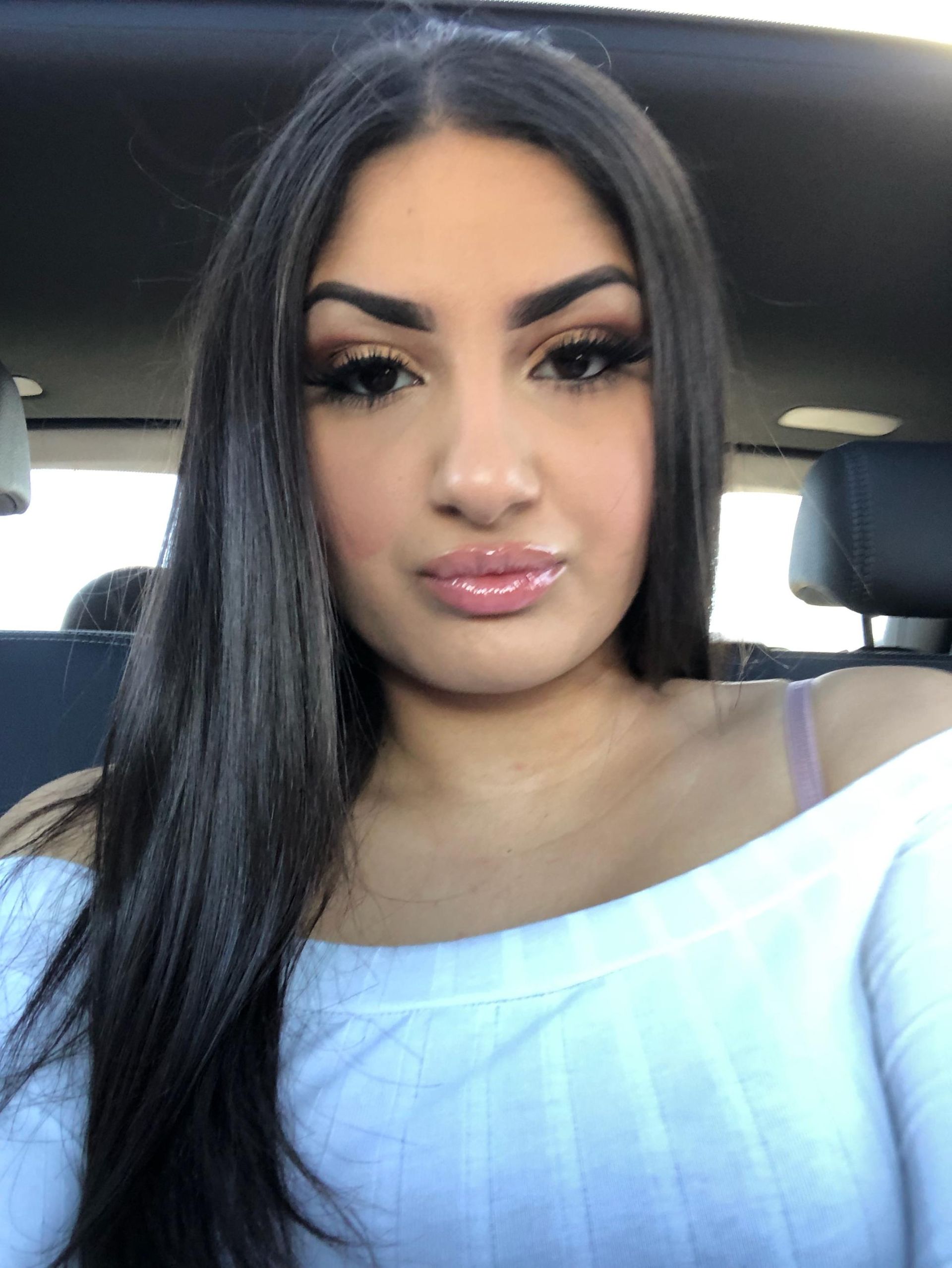Person with long dark hair and makeup, wearing a white off-shoulder top, seated in a car.
