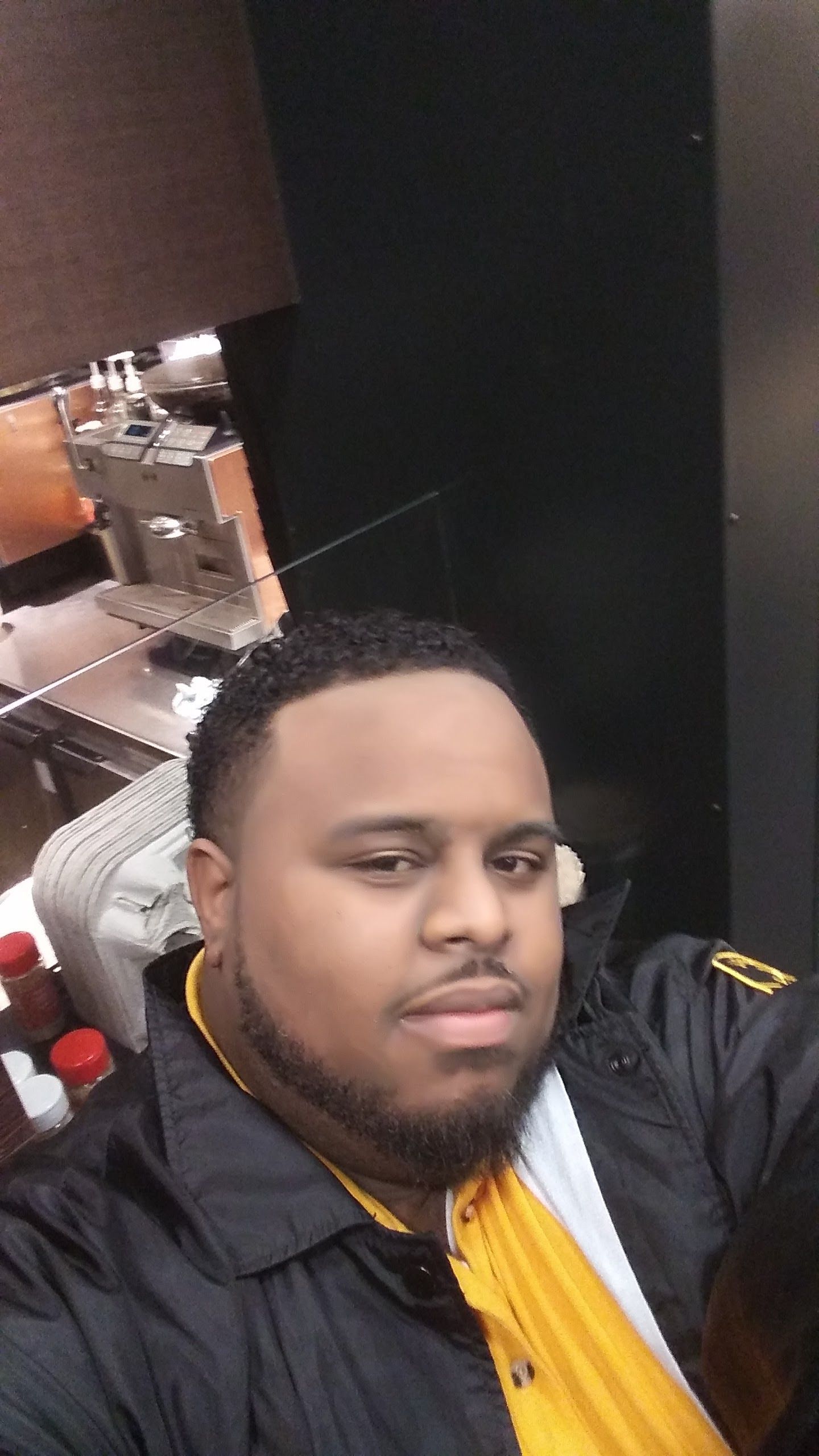 Selfie of a man in a black jacket and yellow shirt, seated indoors near a lamp and table.