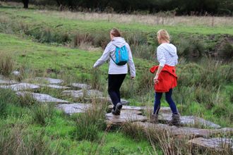 Twee mensen lopen over stapstenen over een grasveld. De een draagt een licht jasje, een rugzak en een zwarte broek.