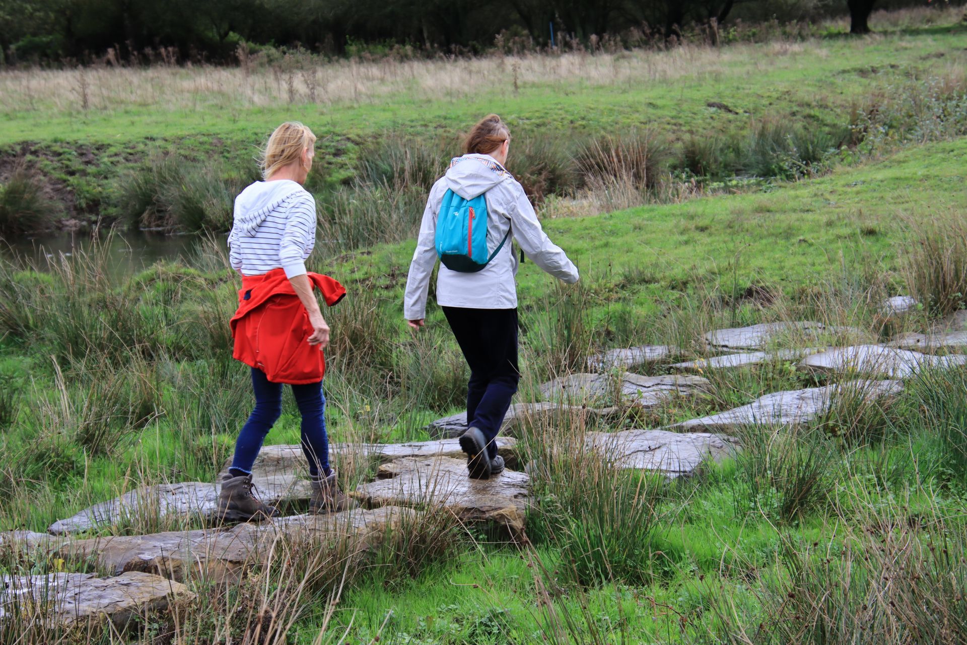 Twee mensen lopen over stapstenen in een grasveld. Een van hen draagt een blauwe rugzak.