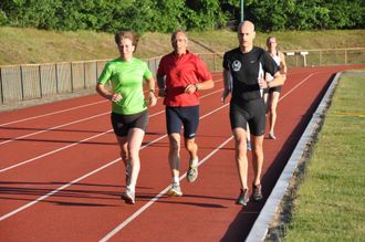 Hardlopers op een baan: vrouw in het groen, man in het rood, man in het zwart en nog een hardloper op de achtergrond.