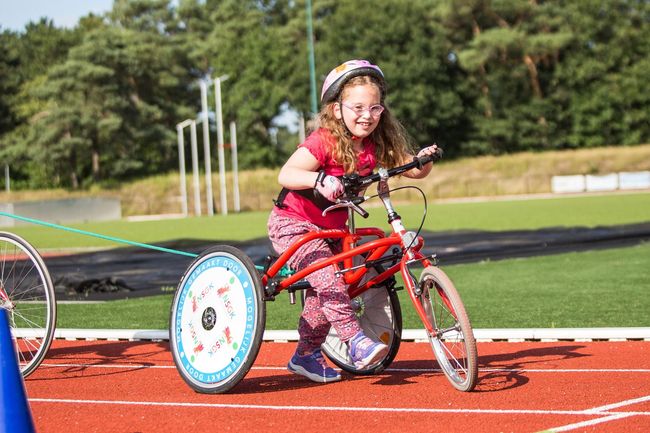 Meisje met bril rijdt lachend op aangepaste fiets op de baan. Rood montuur, helm.