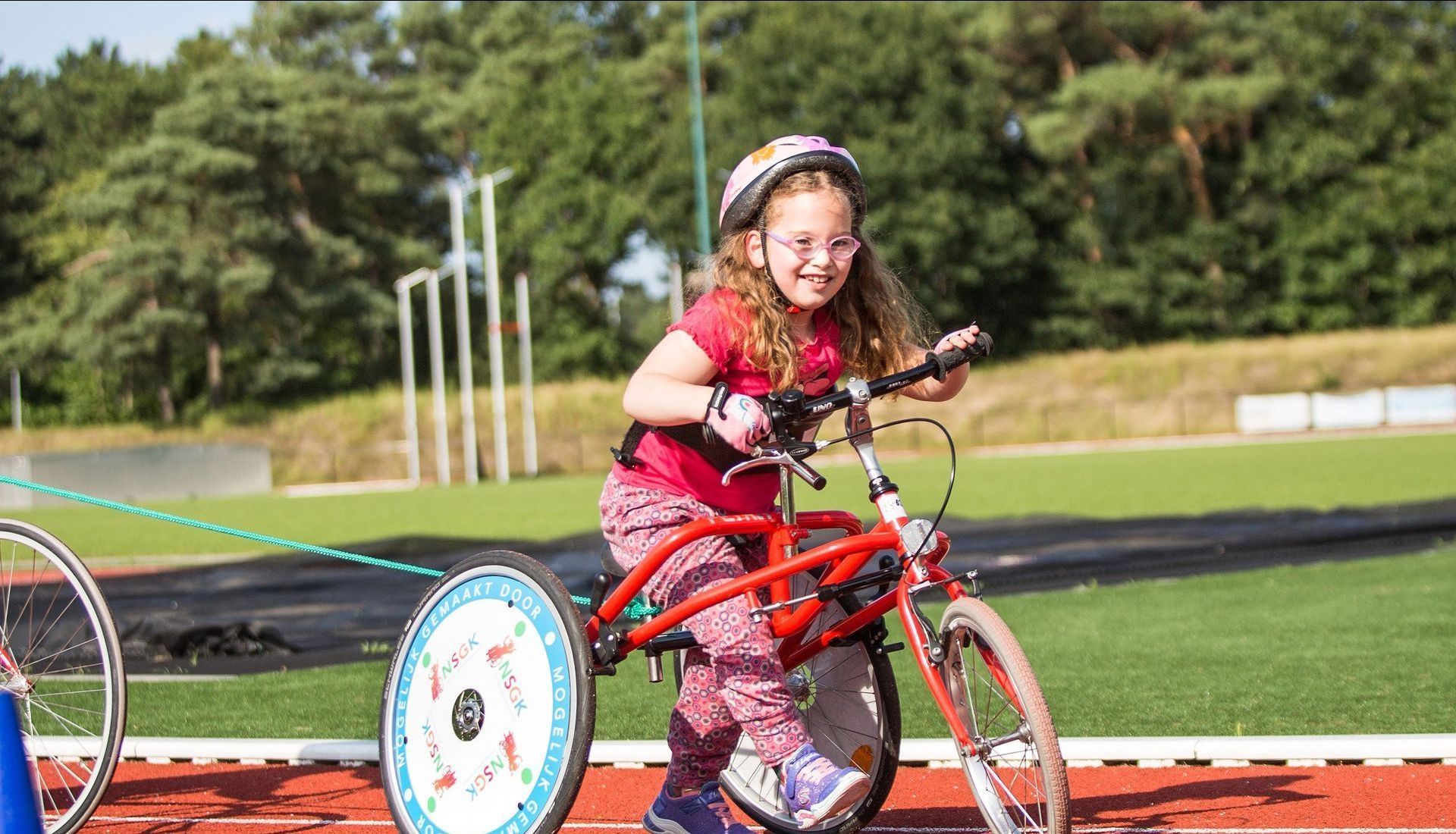 Meisje op een rode aangepaste fiets op een parcours, glimlachend van plezier, met een helm en een roze shirt aan.
