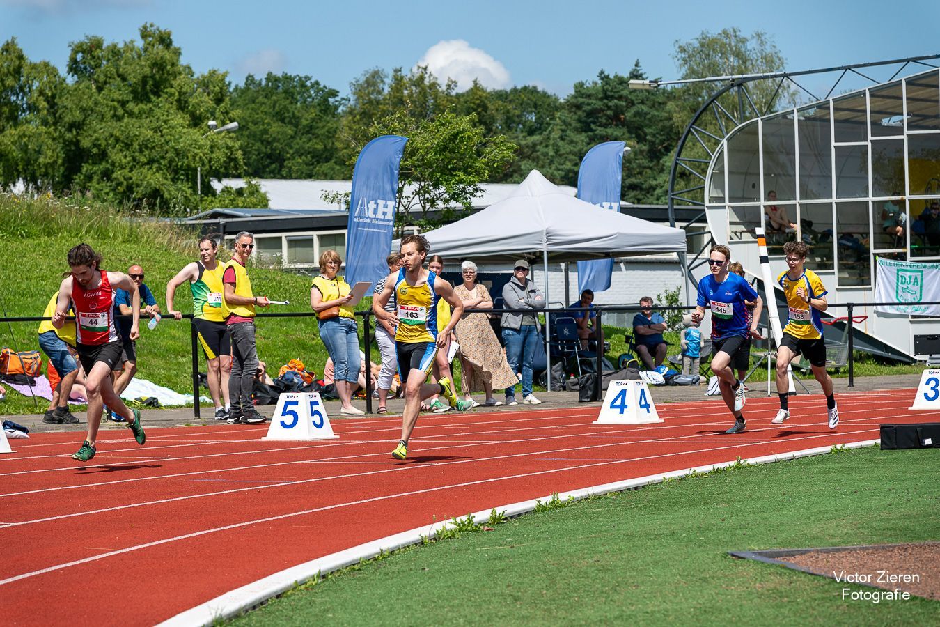 Meisje met bril rijdt lachend op aangepaste fiets op de baan. Rood montuur, helm.