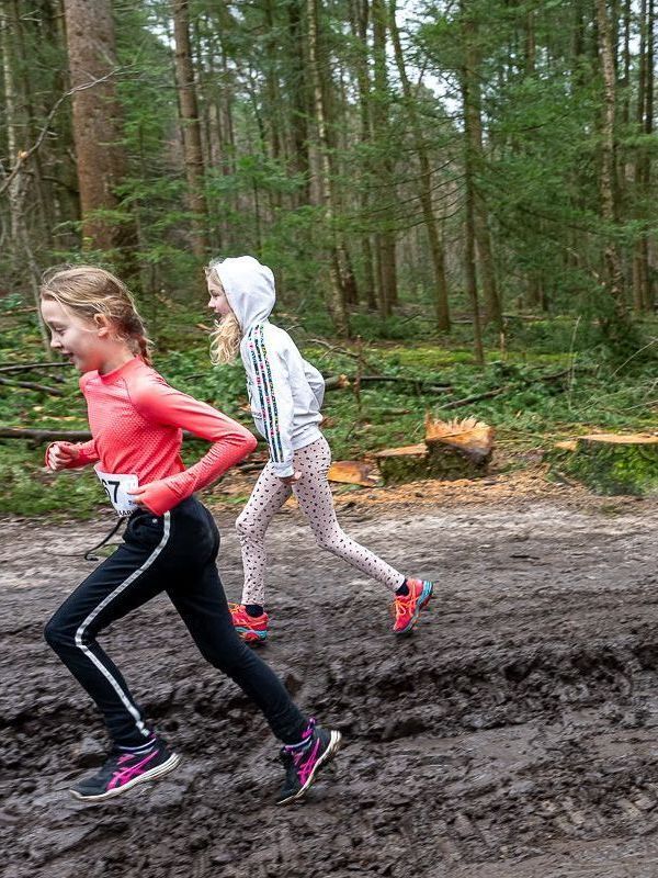 Twee meisjes rennen in sportkleding over een modderig bospad.