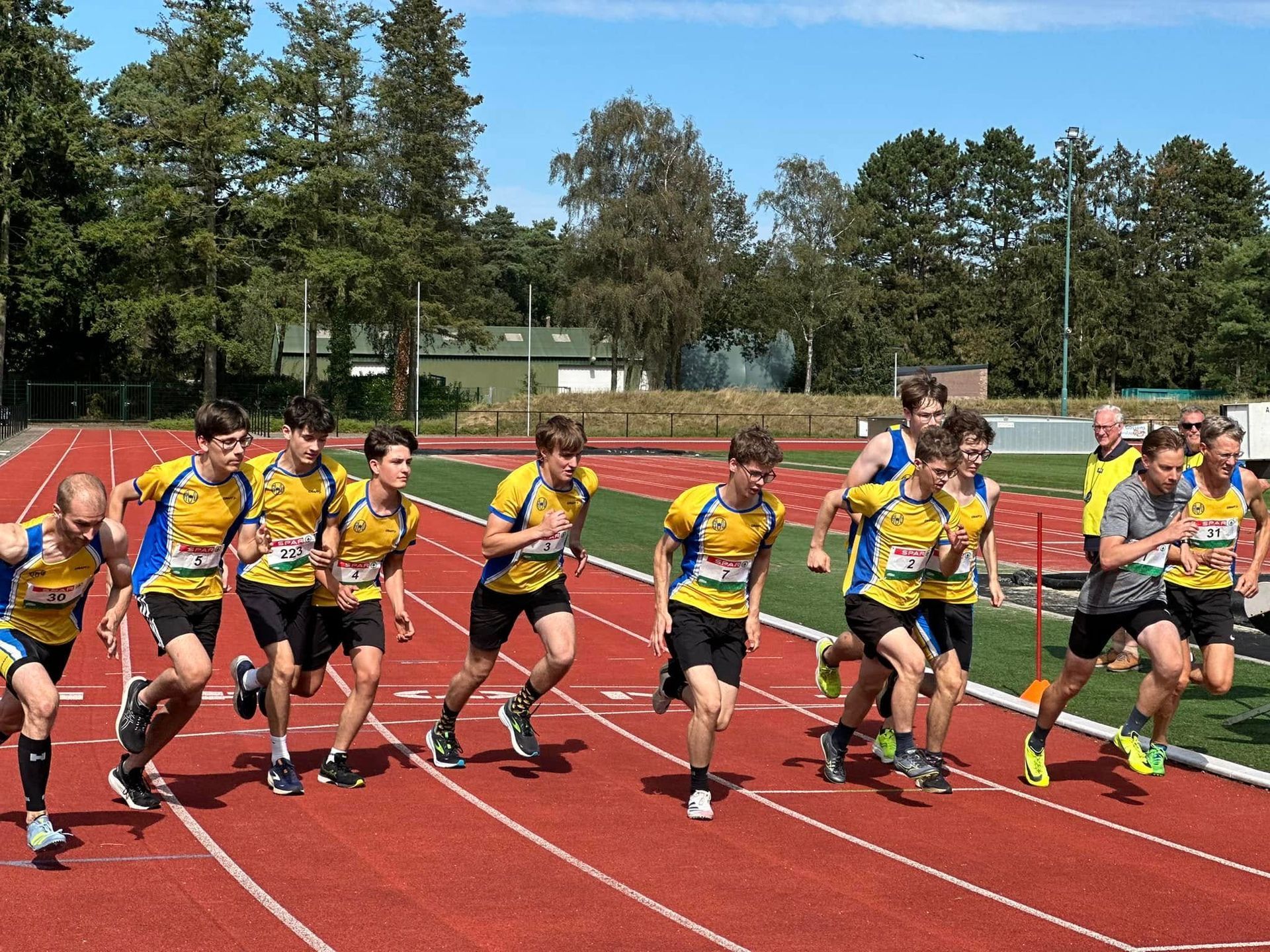 Lopers in geel-zwarte uniformen starten op een rode baan op een zonnige dag.