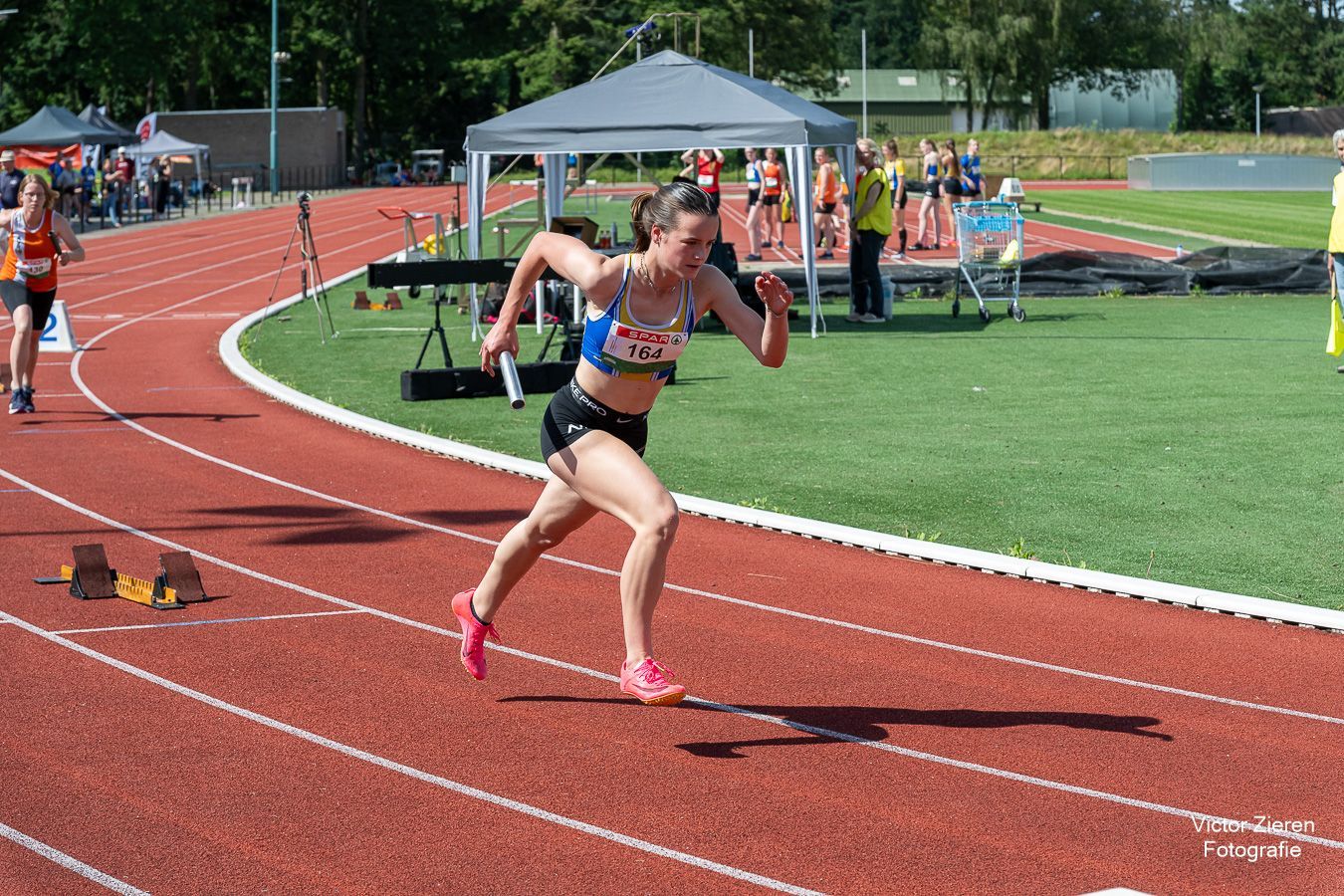 Vrouwelijke atleet rent op een rode baan, met een estafettestokje in haar handen. Het is een zonnige dag.