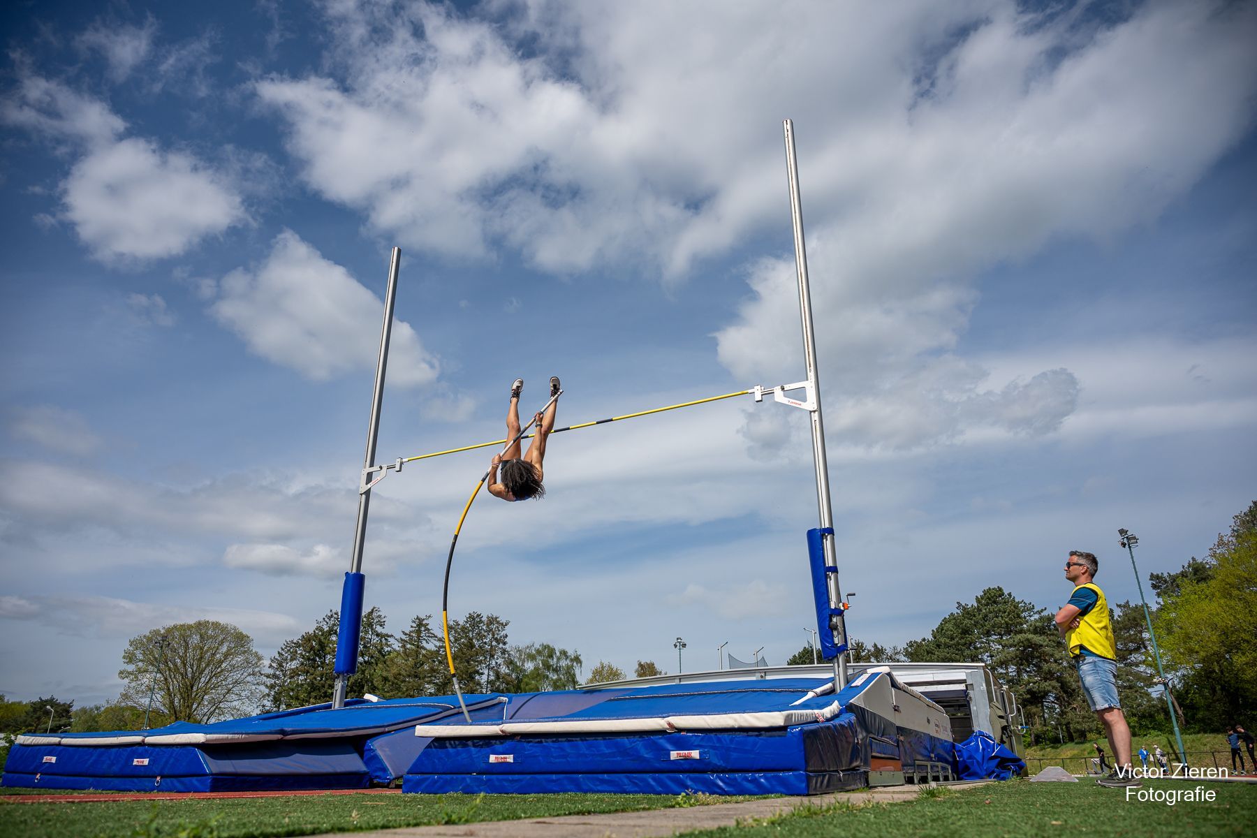 Polsstokhoogspringer in de lucht, over de lat. Blauwe uitrusting, zonnige buitenlocatie.