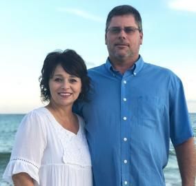 A woman and a man standing close, smiling. They are in front of the ocean. The woman wears white and the man wears blue.