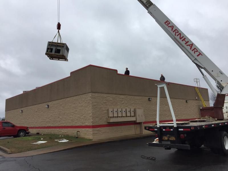 A crane lifts an HVAC unit onto a low commercial building roof. Two workers stand on the roof.