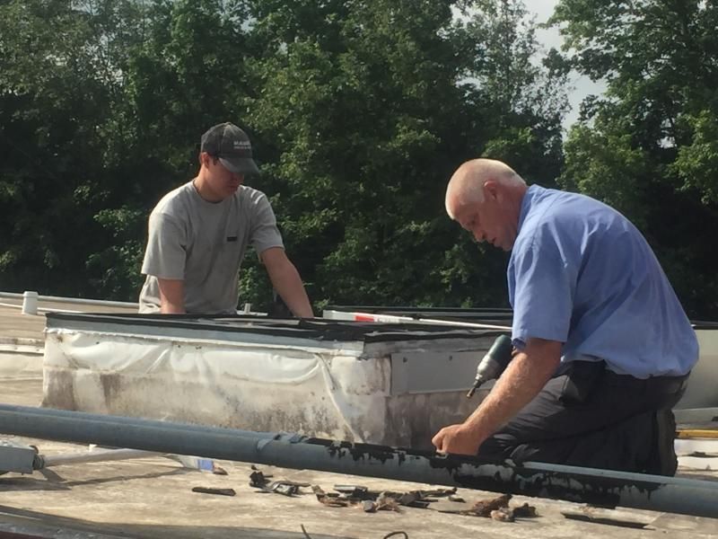 Two people on a rooftop repair a skylight, with tools and materials visible.
