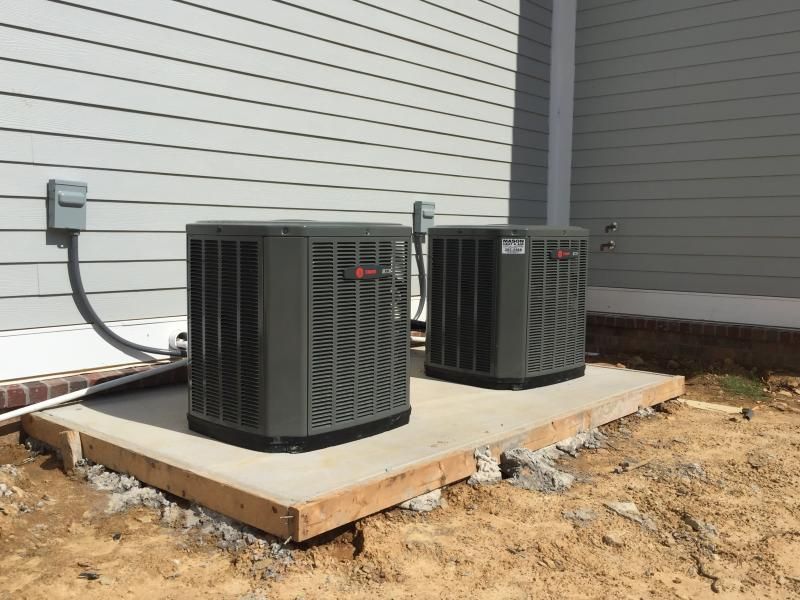 Two dark gray HVAC units on a concrete pad next to a light gray house.