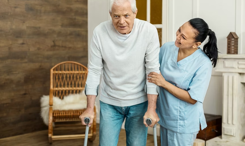 Man using crutches, assisted by a person in a blue scrub top. They are inside a room with wood paneling and a rocking chair.