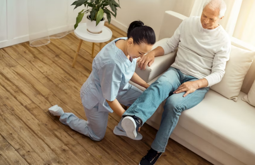 Woman in scrubs assists a man, examining his leg. They are in a well-lit living room.