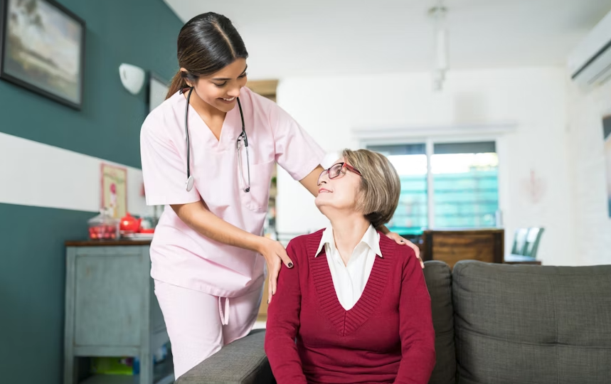 Caregiver in pink scrubs assists an older person in a red sweater sitting on a couch.