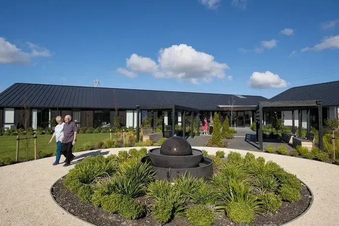 Couple walking towards a modern building with a fountain and garden on a sunny day.