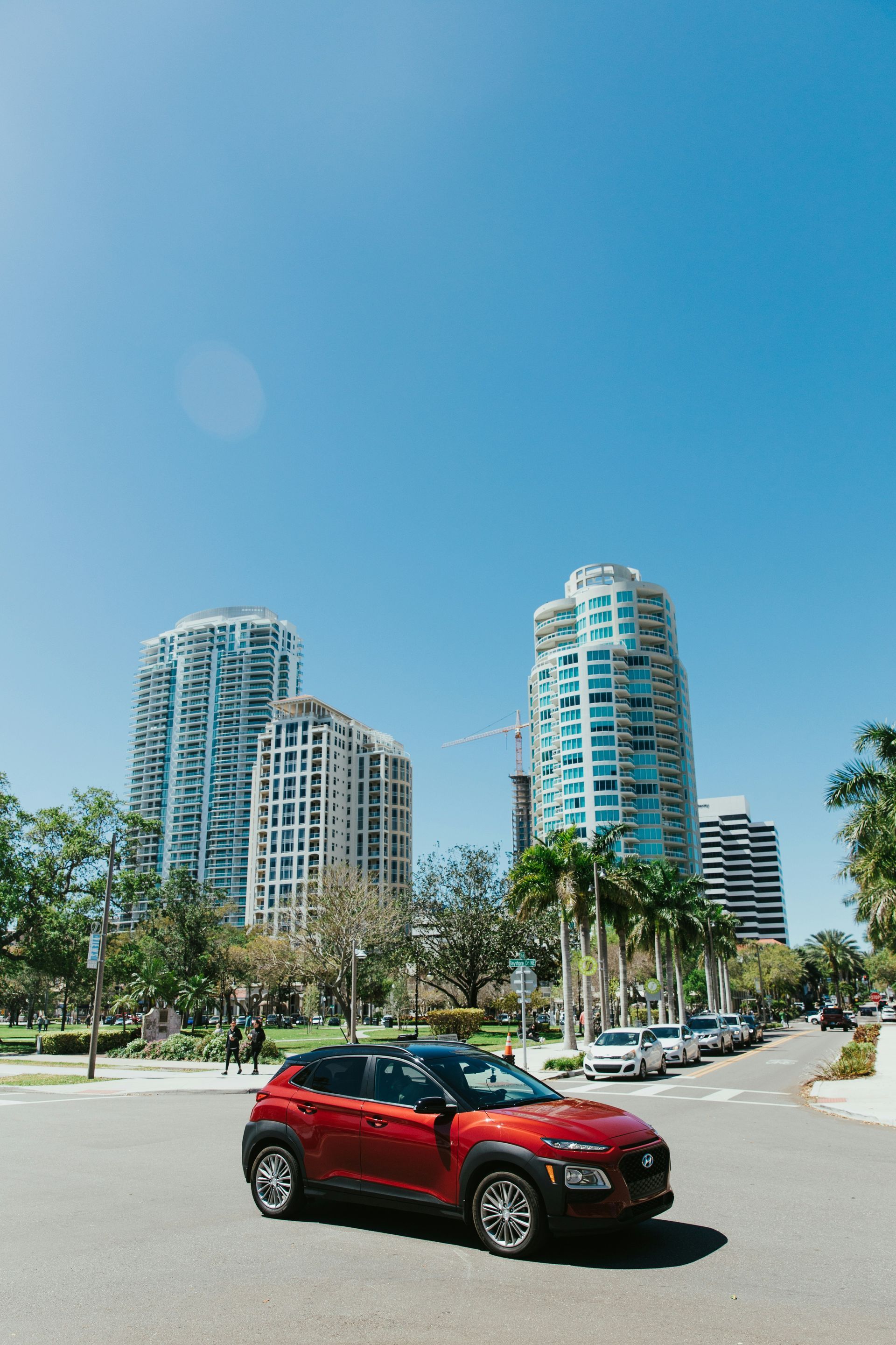 A red car is driving down a city street with tall buildings in the background.