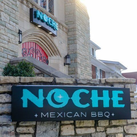 A sign for noche mexican bbq is on a stone wall