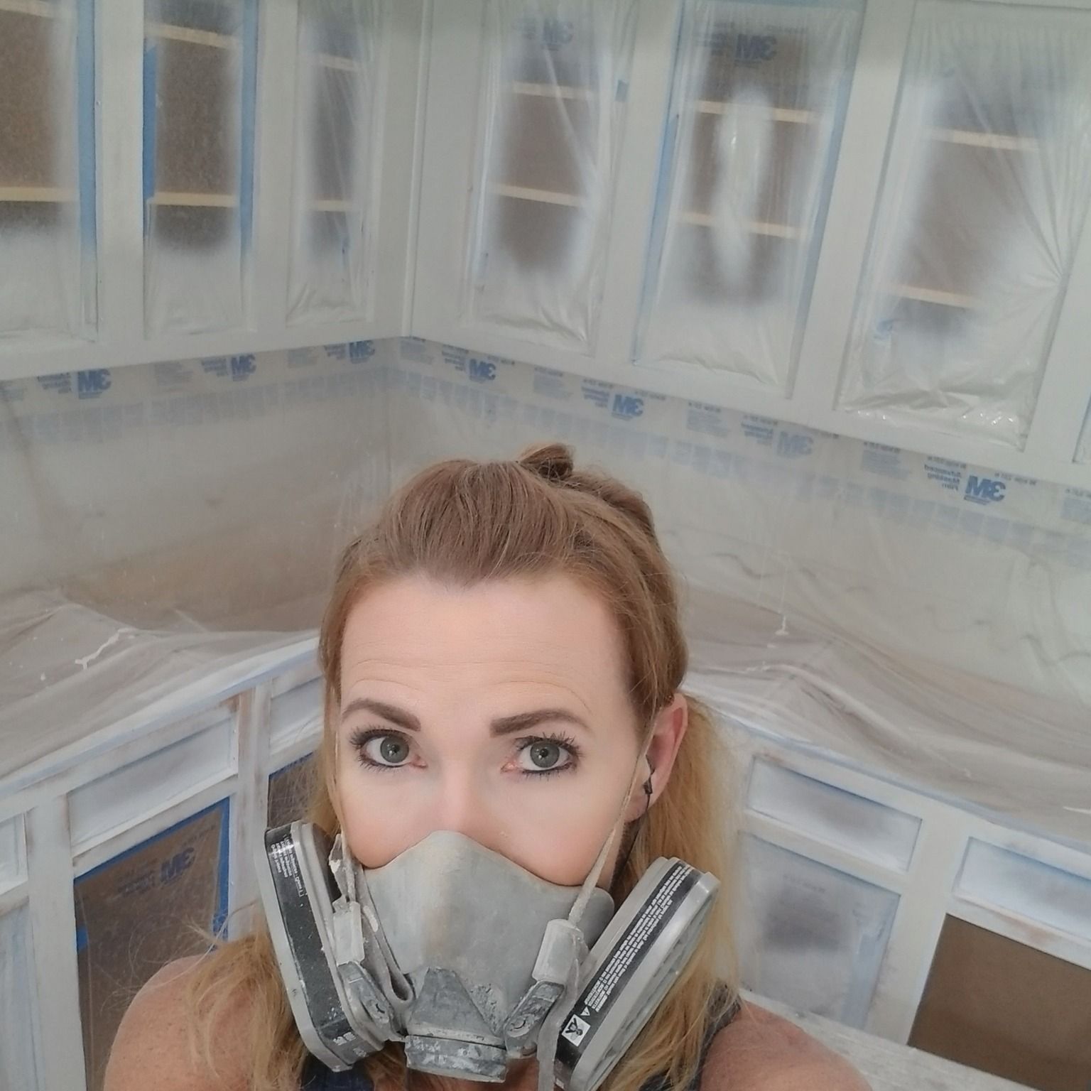 Woman in respirator painting kitchen cabinets white, covered in plastic.