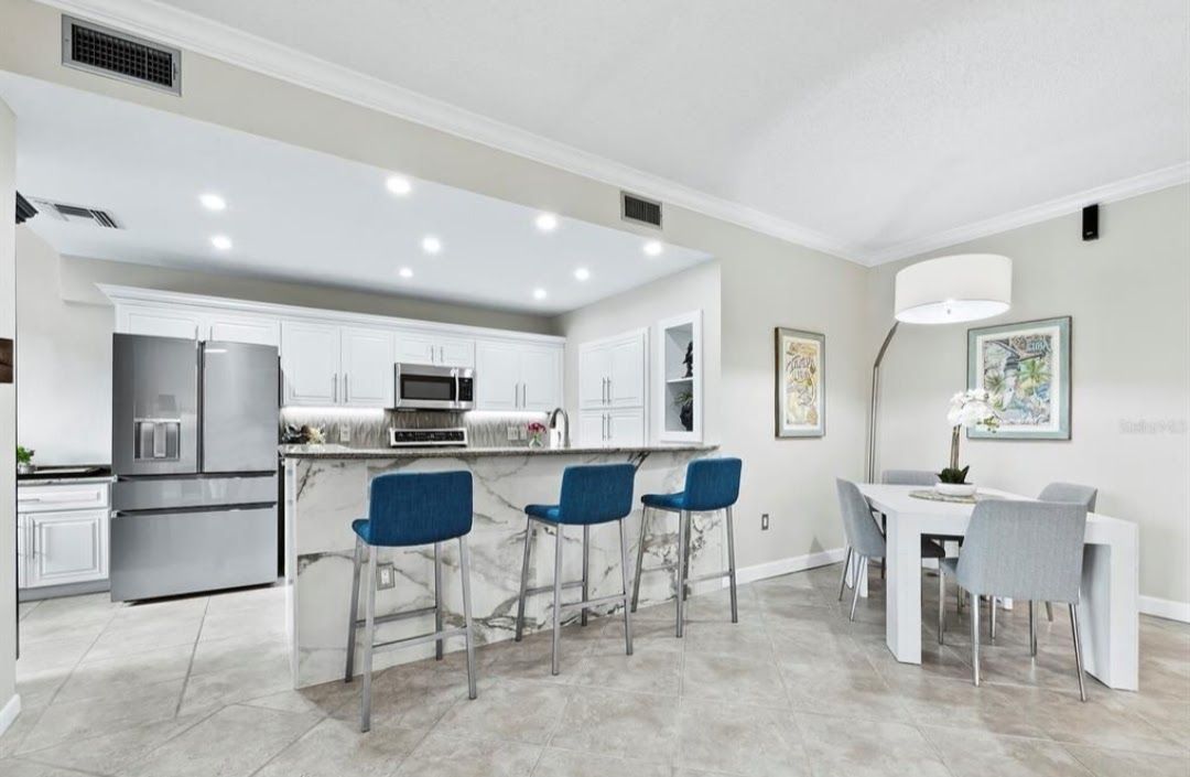 Open-concept kitchen with marble island, stainless steel appliances, blue bar stools, and dining area with white table and gray chairs.