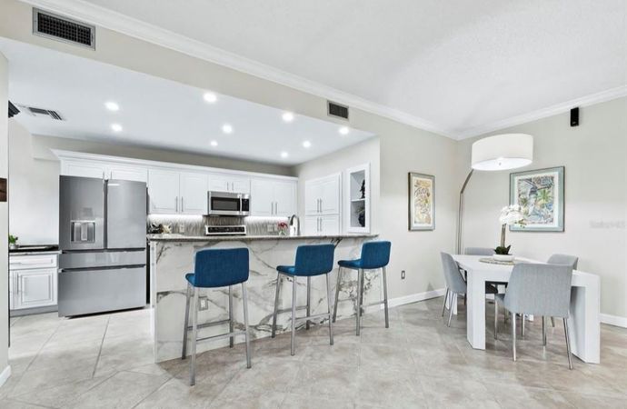 Open-concept kitchen with marble island, stainless steel appliances, blue bar stools, and dining area with white table and gray chairs. Open-concept kitchen with marble island, stainless steel appliances, blue bar stools, and dining area with white table and gray chairs.