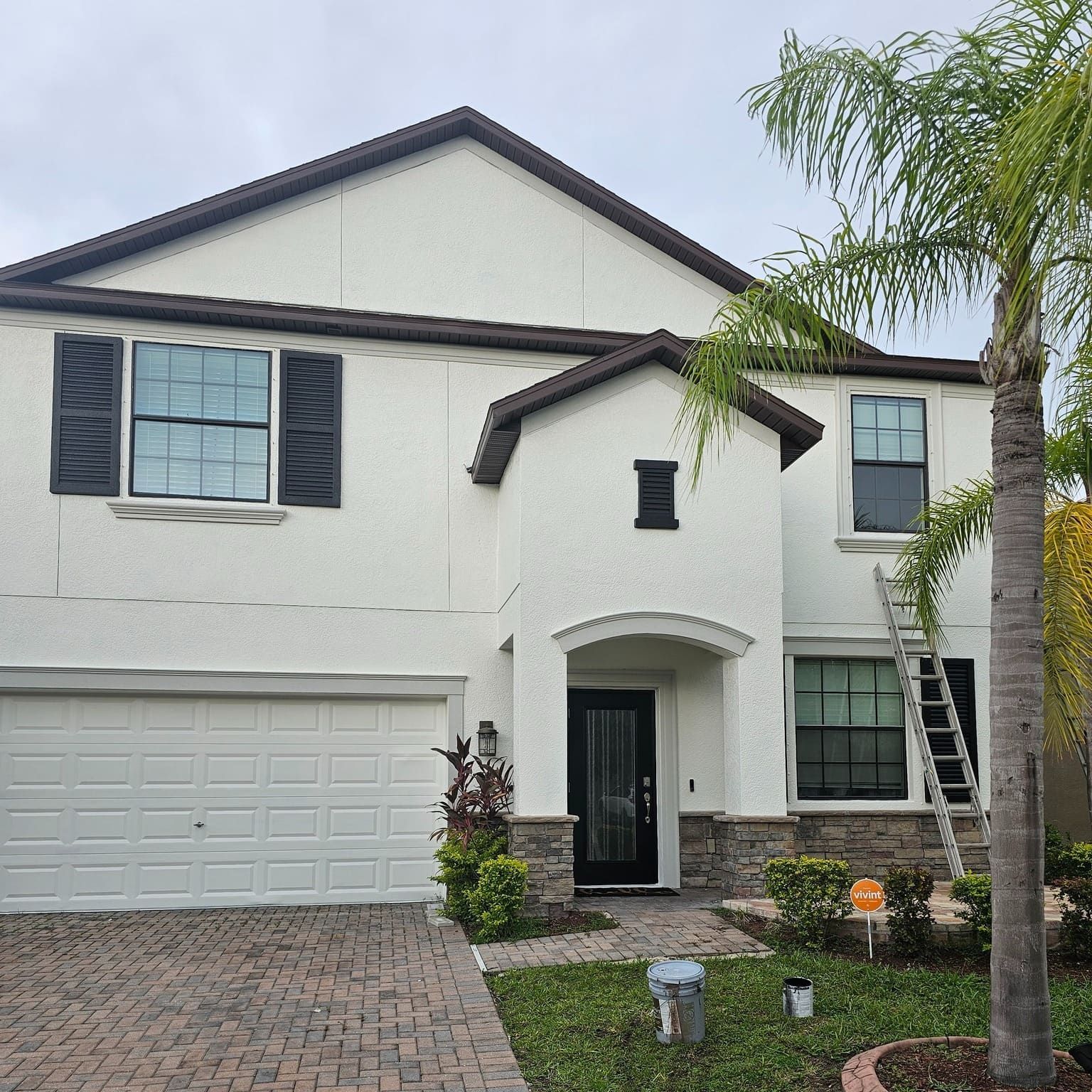 Two-story white house with brown accents and shutters. A ladder leans against the side, palm tree in view. Two-story white house with brown accents and shutters. A ladder leans against the side, palm tree in view.