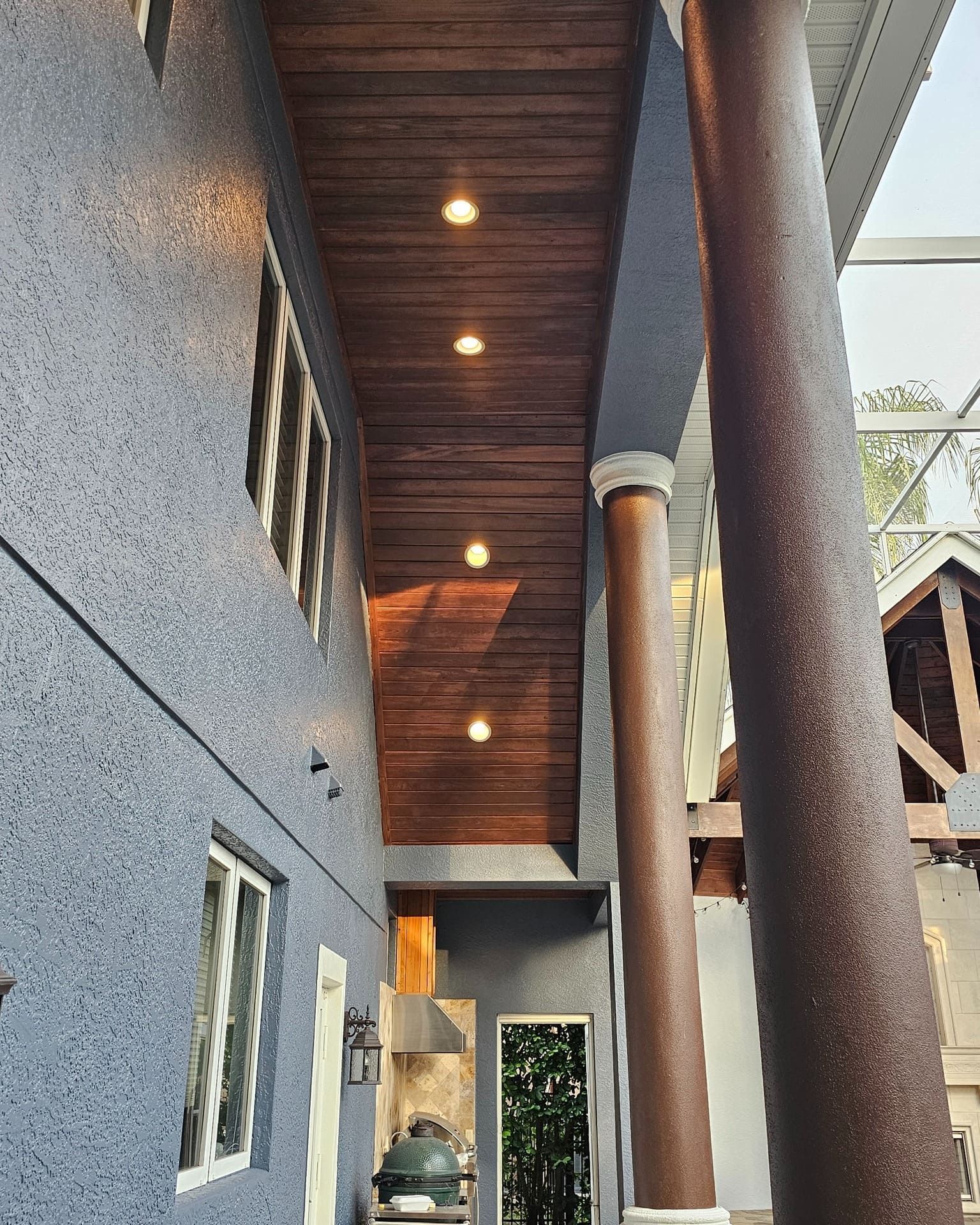 Dark wood ceiling with recessed lights, blue textured wall, brown columns, outdoor setting. Dark wood ceiling with recessed lights, blue textured wall, brown columns, outdoor setting.