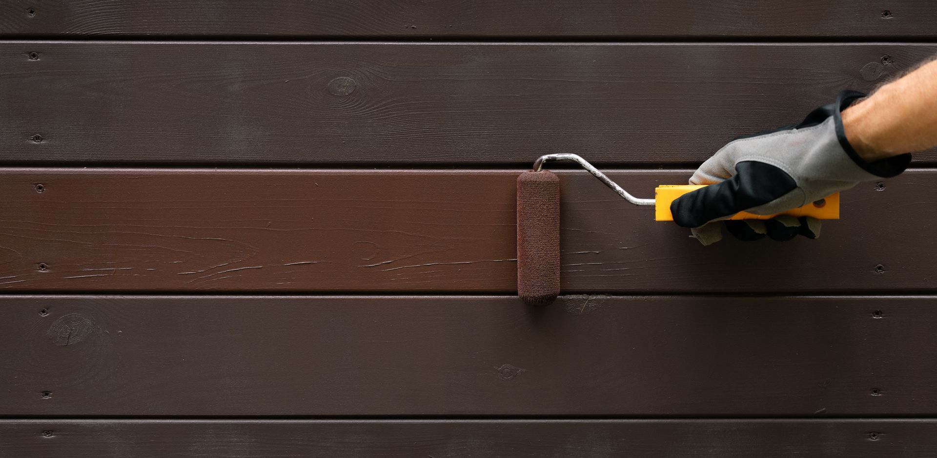 Hand with paint roller painting wooden house exterior.