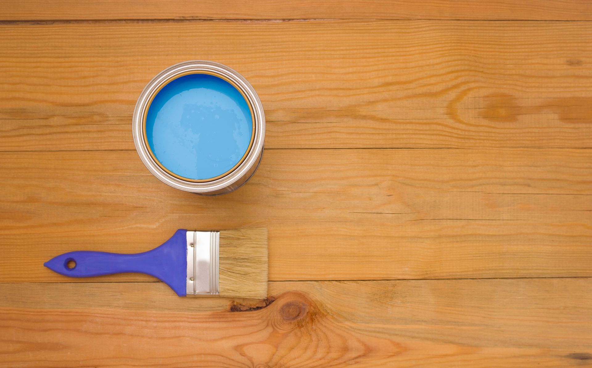 Open blue paint can and brush rest on warm wood surface.