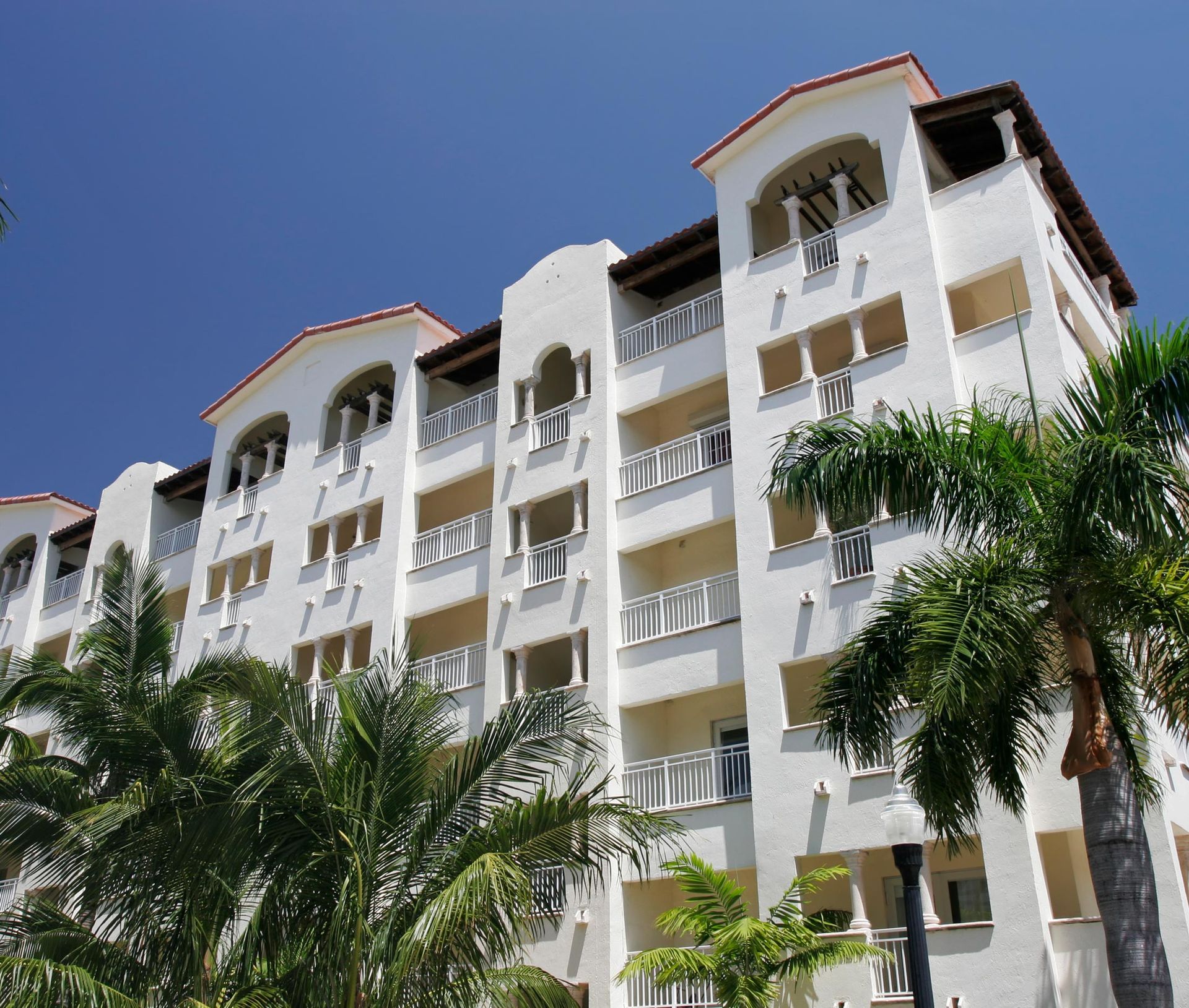 White apartment building with balconies and palm trees against a blue sky.
