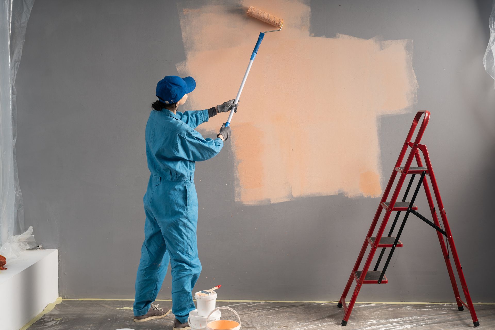 Person in blue coveralls paints a wall with an orange roller, red ladder in the room.