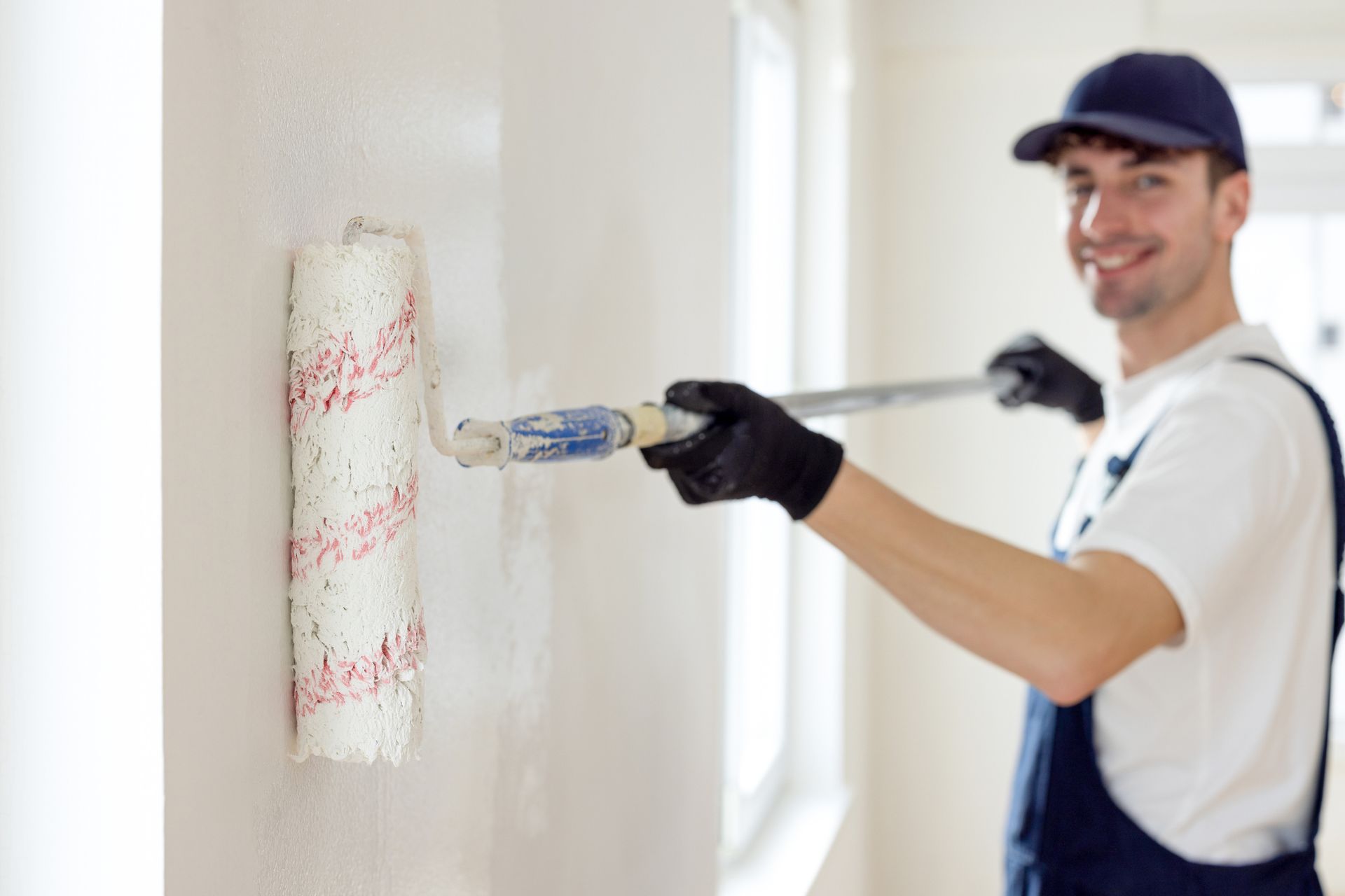Man wearing overalls painting a bright room with a roller brush.