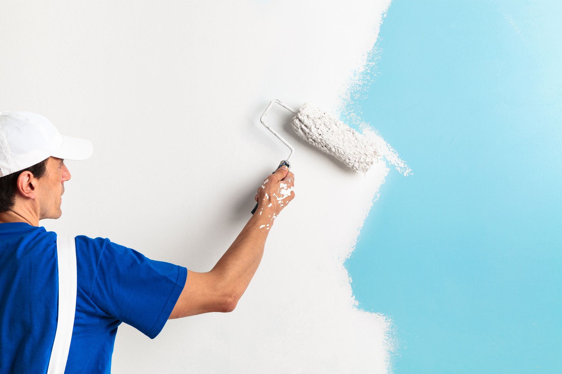 Man in blue shirt and white cap painting a wall light blue with a roller.