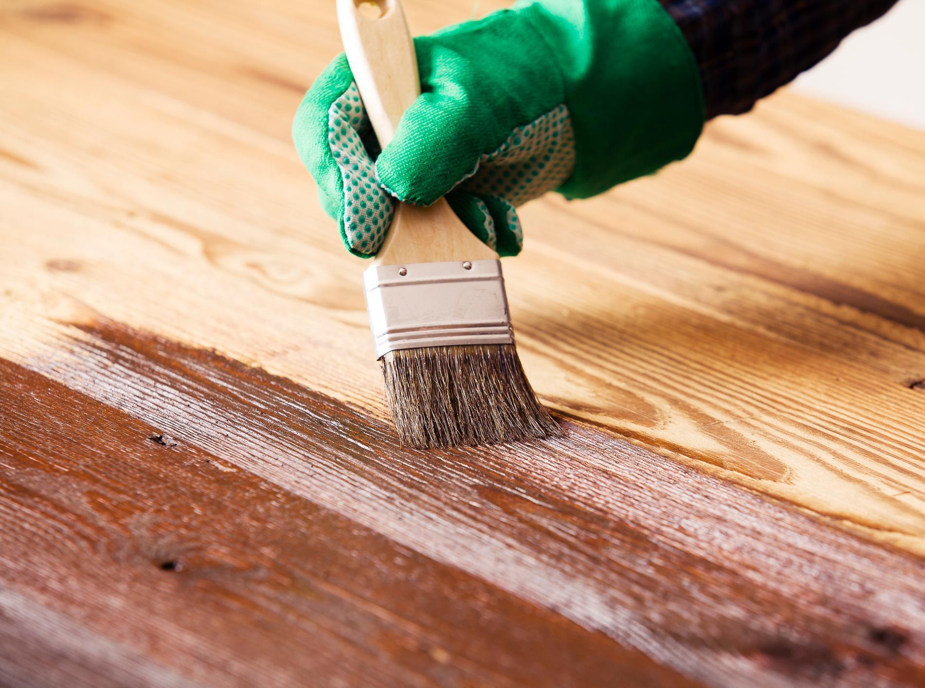 Hand wearing a green glove applying wood stain with a brush to a wooden surface. Hand wearing a green glove applying wood stain with a brush to a wooden surface.
