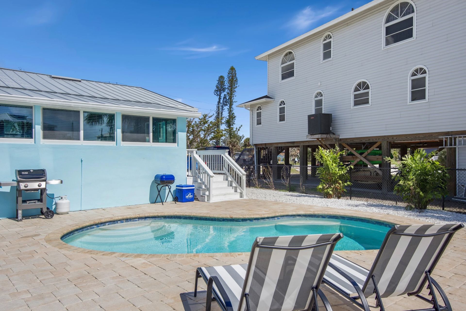 A pool deck with two striped lounge chairs overlooks a curved swimming pool next to a light blue building and stilt house.