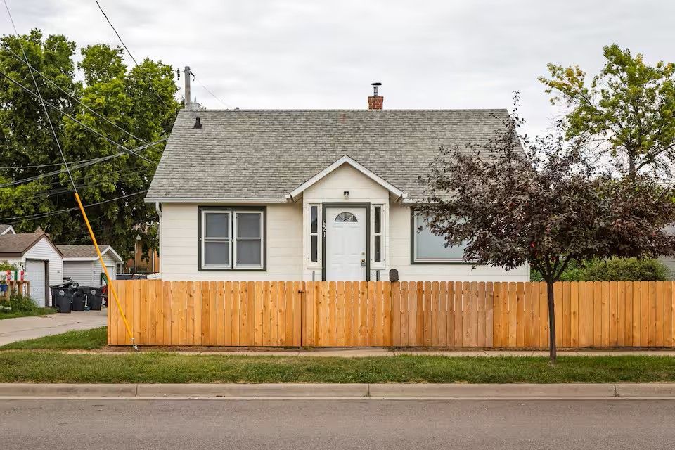 A single-story, light-colored house with a peaked roof and a tall, wooden picket fence along the front yard.